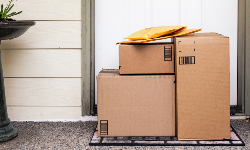 Three cardboard packages and two yellow padded envelopes are stacked on a doorstep beside a white door, sitting on a welcome mat. An atmosphere of anticipation.