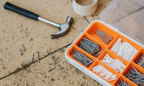 A hammer lies on a concrete surface surrounded by scattered nails. An open orange toolbox displays organized screws and wall plugs. A roll of tape is beside it.